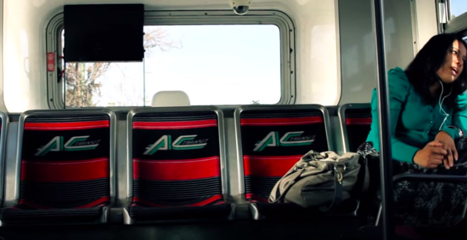 Image of a person sitting on a bus looking out the window mid song. To the person's left are three empty bus seats. Behind the seats is a window.
