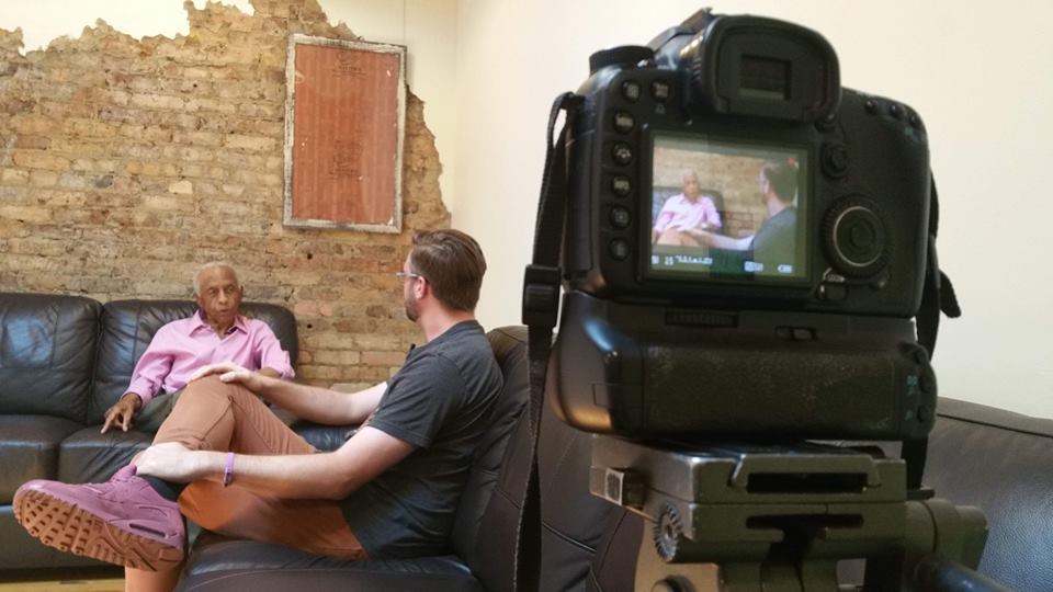 a camera in the foreground records a man interviewing an older man about his experiences